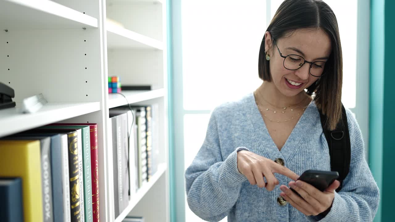 年轻美丽的西班牙女学生在图书馆大学用智能手机找书视频素材