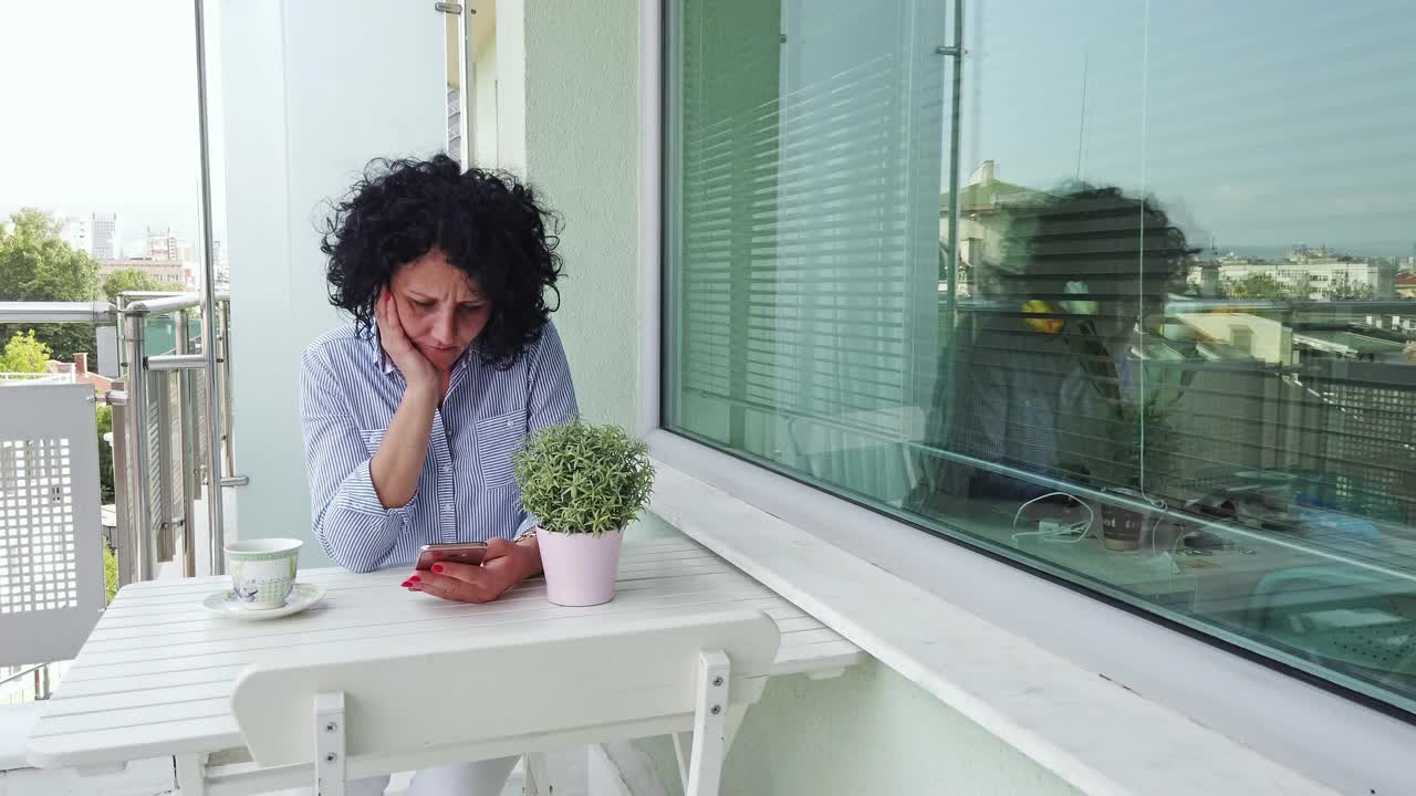 快乐的西班牙女人喝咖啡和使用手机坐在露台上视频素材