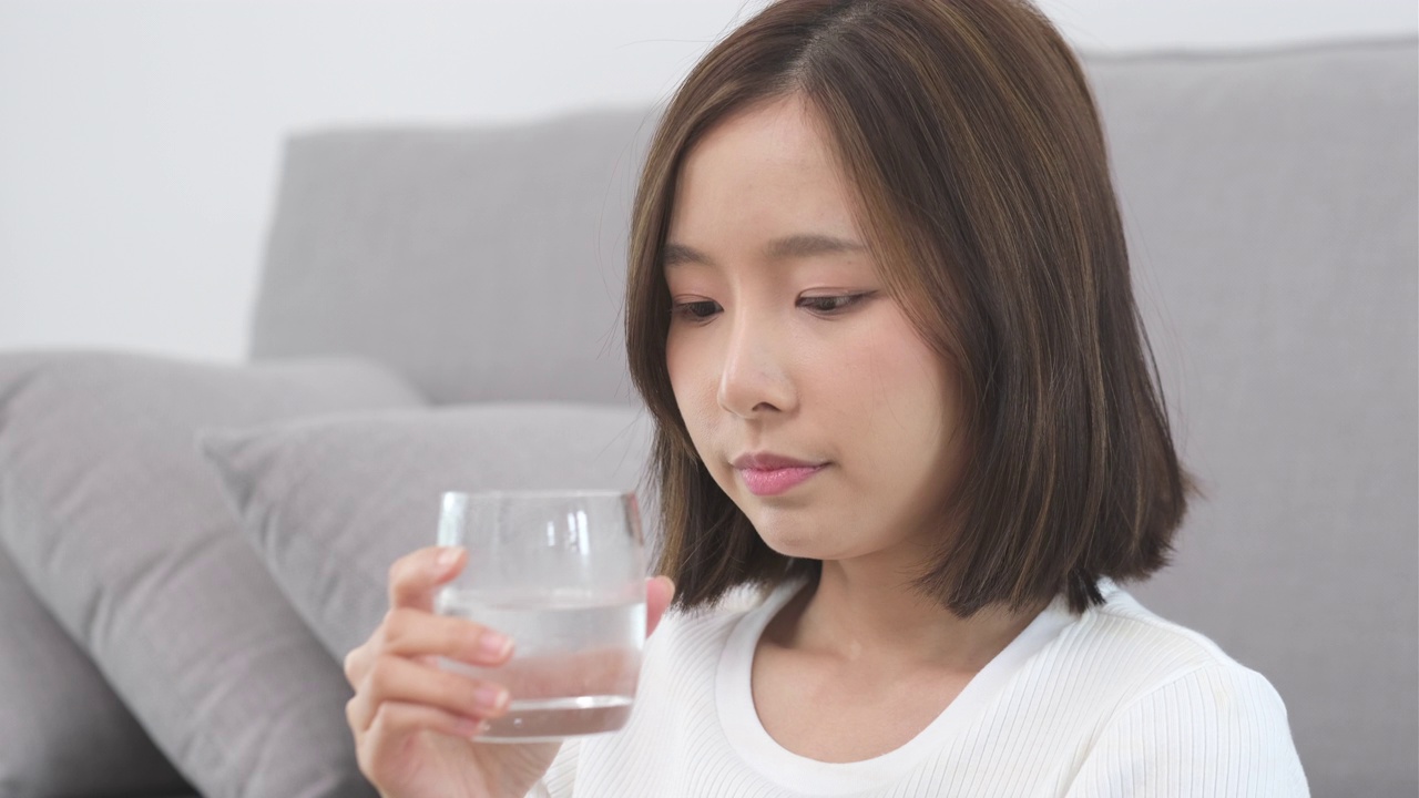 年轻的亚洲女子坐在家里的客厅里喝淡水视频素材