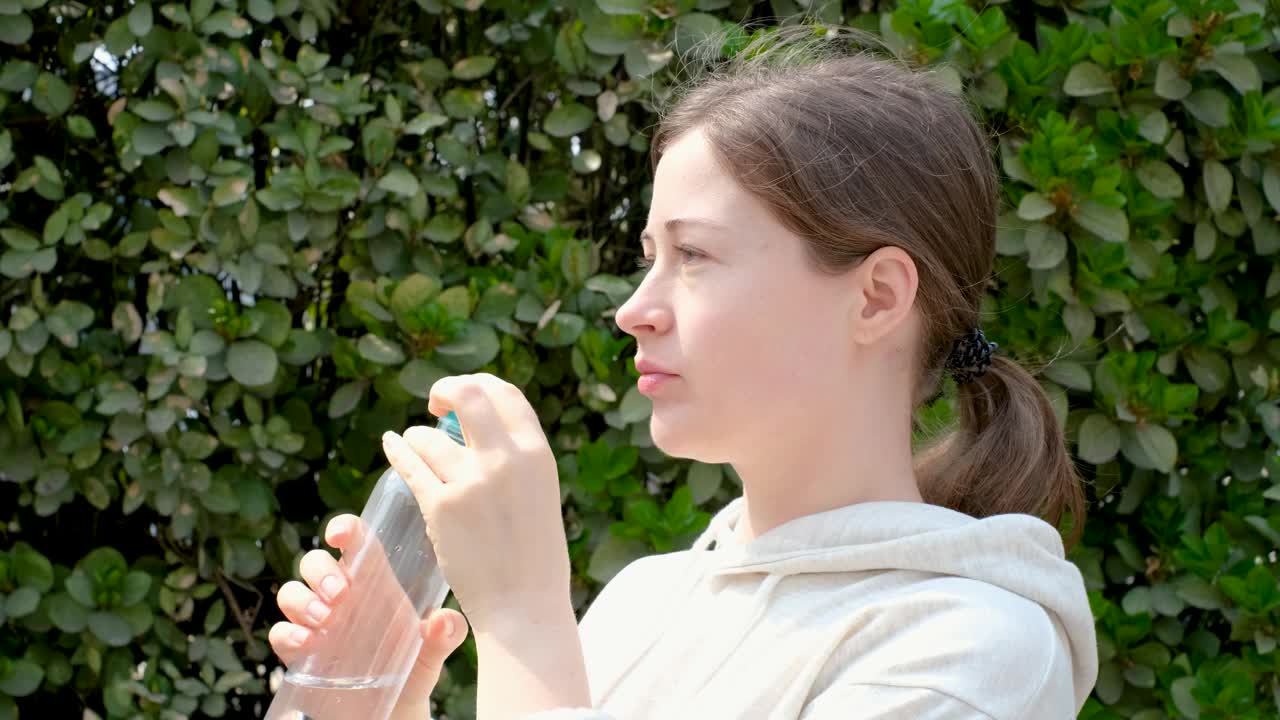 一个年轻女子的肖像从塑料瓶喝水在绿色的叶子墙的背景。视频素材