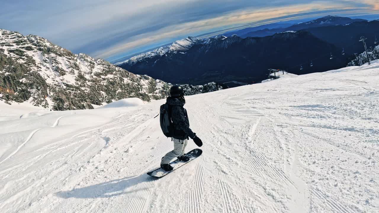 在阳光下，滑雪板运动员从山坡上骑下来，踩着刚下的雪粉。一座美丽的山展现在骑手的面前，天空中也有云。股票视频视频素材