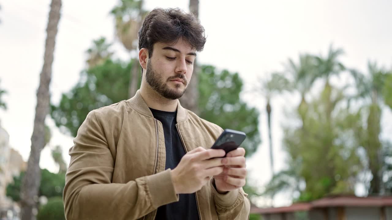 年轻的西班牙裔男子在公园用智能手机微笑视频素材