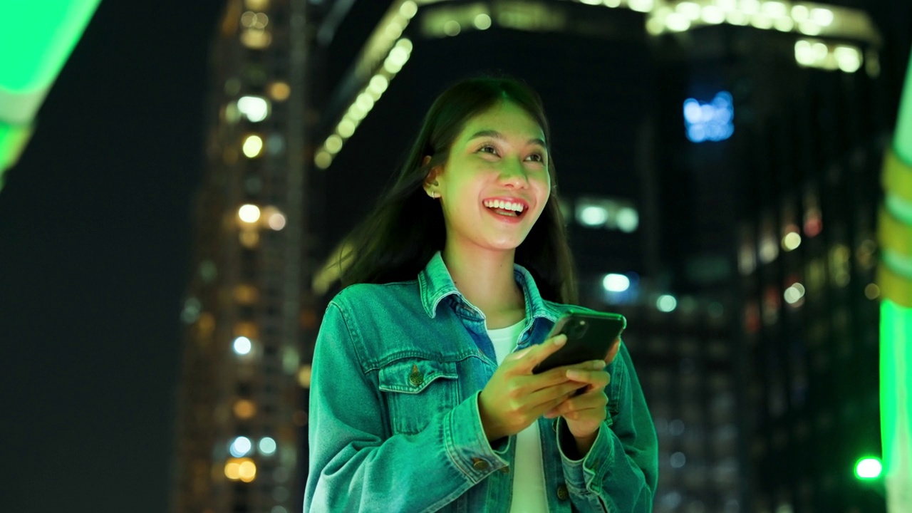 美丽的年轻亚洲女子在晚上用手机走在城市街道上。一个快乐的女人站在那里用手机打字视频素材