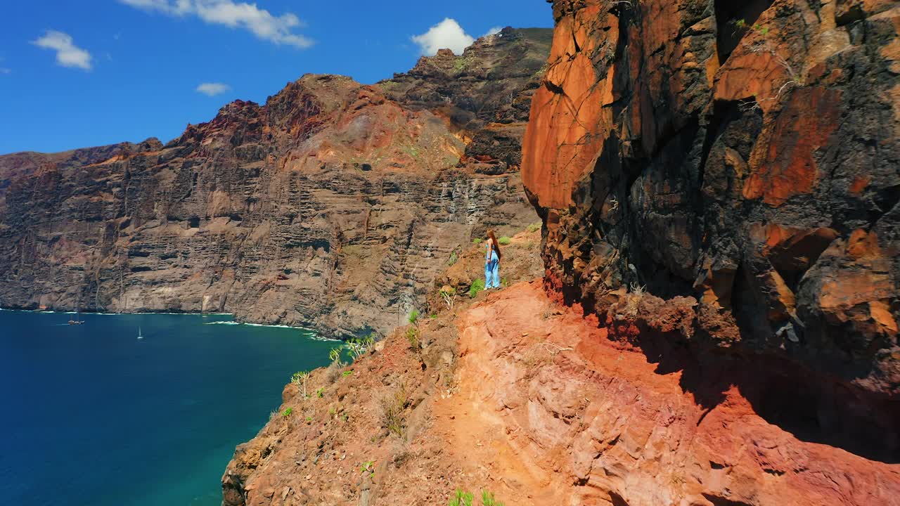 游客女孩的后视图剪影站在火山岩的边缘，享受海景与令人难以置信的巨大洛斯吉甘蒂斯悬崖加那利群岛特内里费。阳光明媚的一天。去西班牙欧洲旅游。视频素材