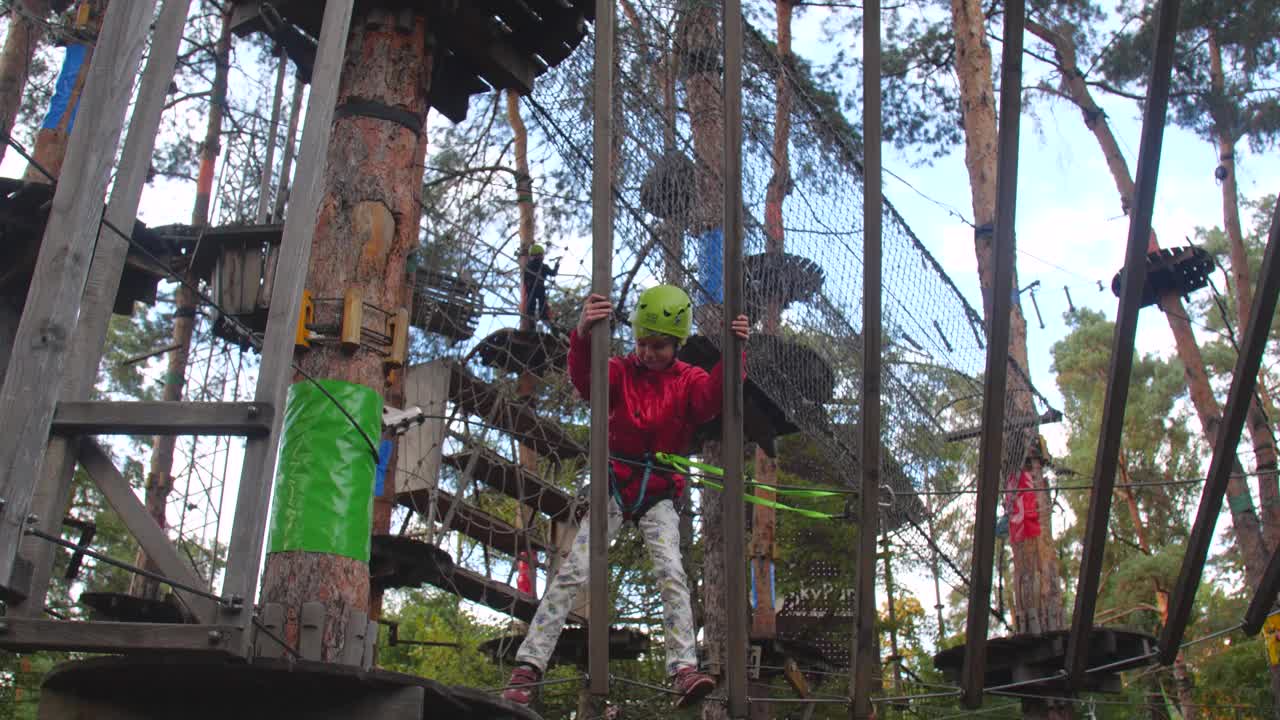 孩子们在绳索公园进行登山训练。一个女孩戴着绿色的头盔，穿着红色的夹克，带着登山装备爬进树林里。儿童运动，积极娱乐视频下载