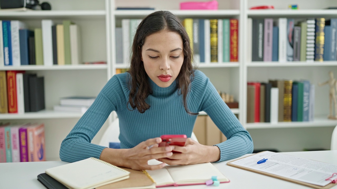 年轻漂亮的西班牙女学生在大学教室里用智能手机在笔记本上写字视频素材