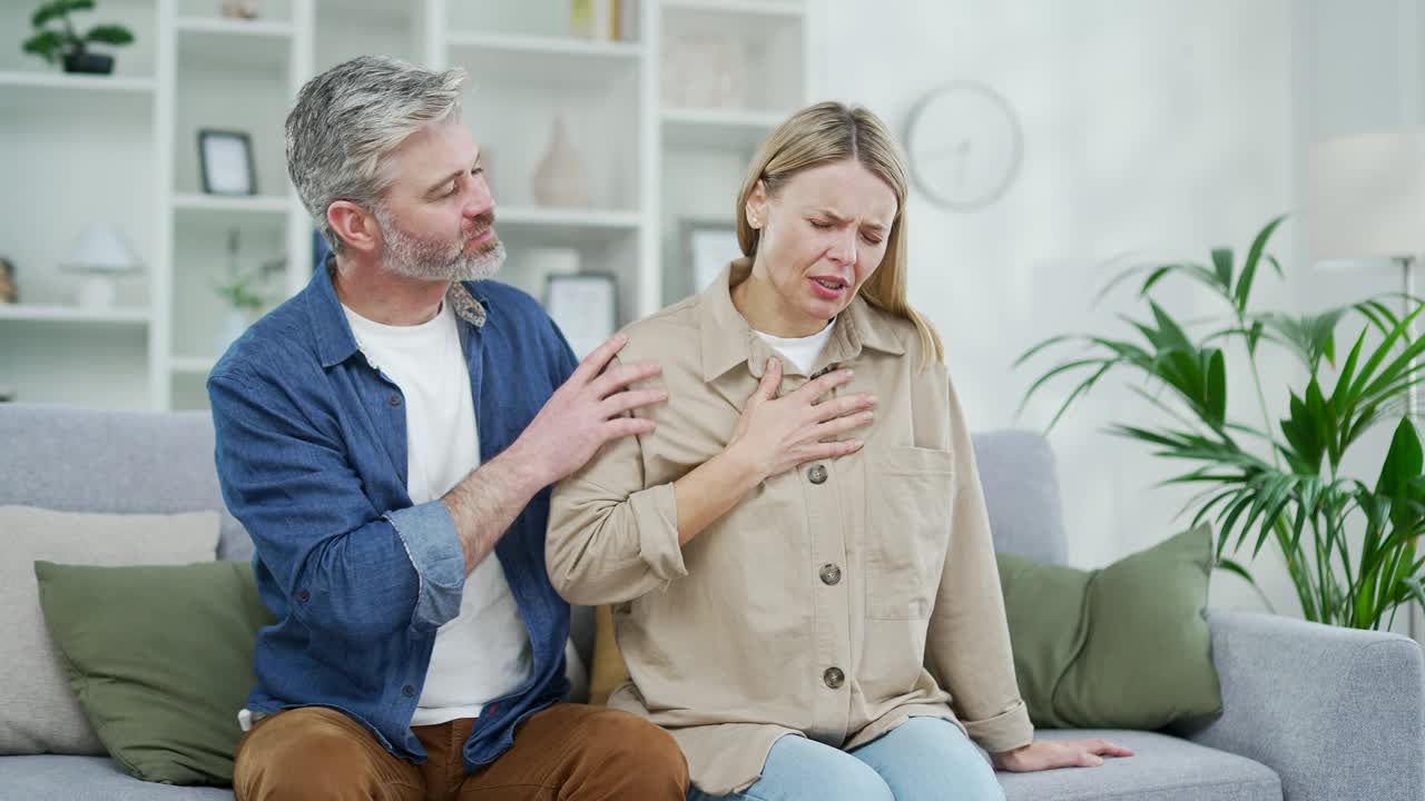 生病的成熟女人坐在家里客厅的沙发上感到胸部紧张性疼痛。丈夫支持妻子，让妻子放心。视频素材