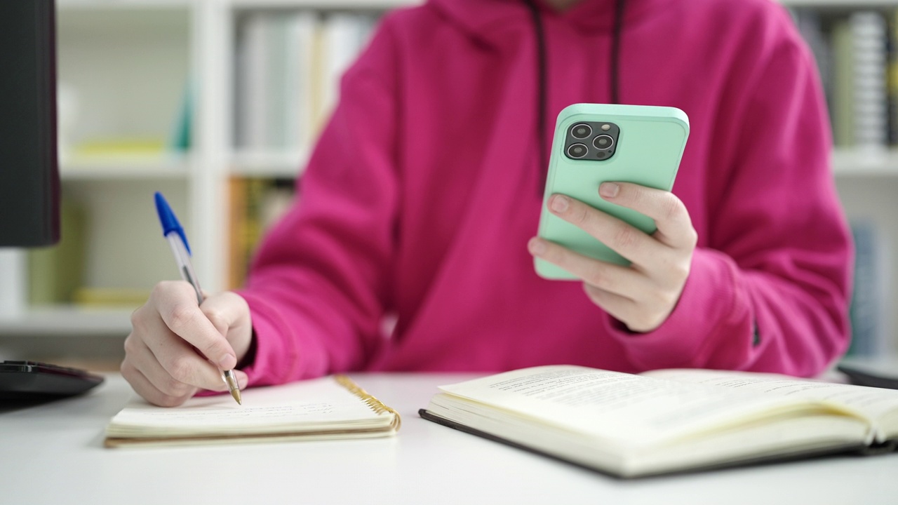 图书馆大学的年轻金发女学生用智能手机在笔记本上写字视频素材