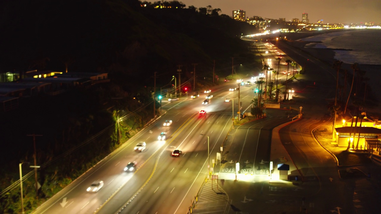 航拍:夜间海上道路上的车辆对着天空倾斜拍摄，无人机向前飞向城市中的照明建筑-加利福尼亚州圣莫尼卡视频素材