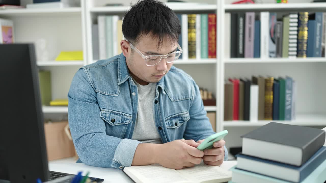 在图书馆大学，年轻的中国男学生坐在桌子上玩智能手机视频素材