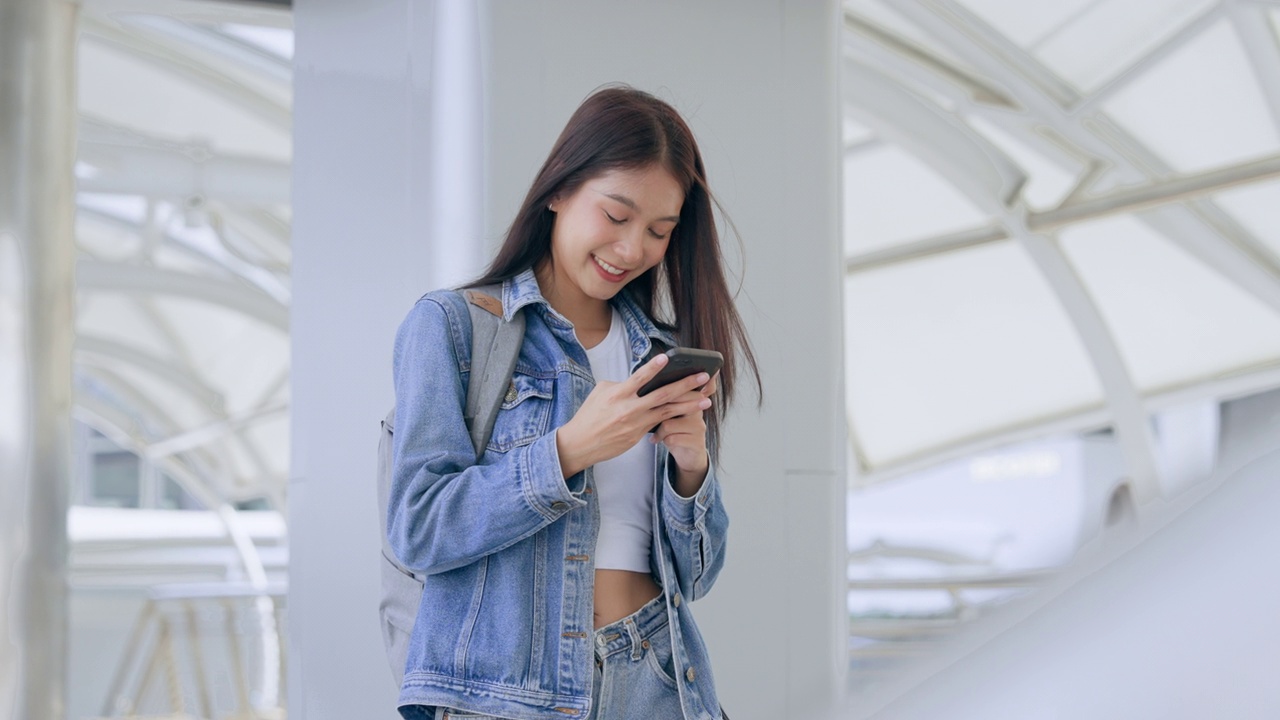 年轻的亚洲女子在户外微笑着使用手机。快乐的女游客穿着牛仔裤夹克，拿着智能手机出现在公共场合视频素材