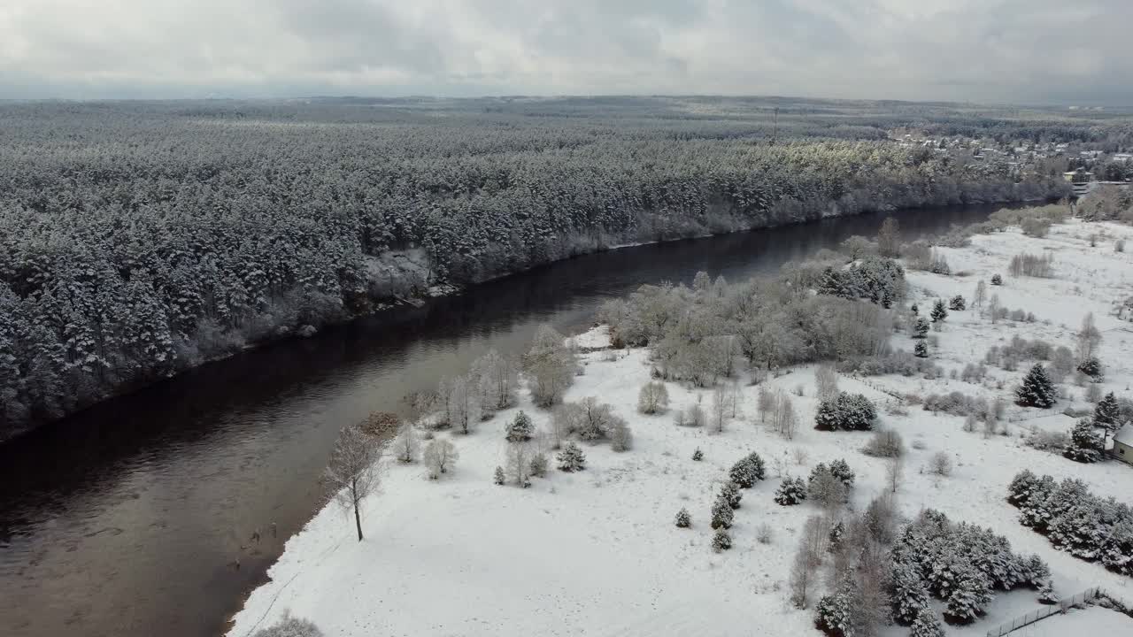 无人机镜头在多云的天空下，在地平线上飞过河边的雪林树木视频素材