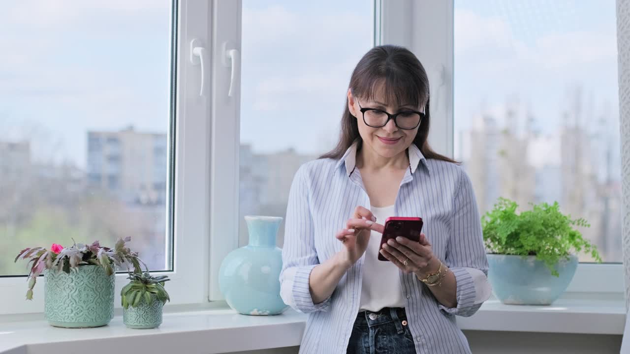 一名中年妇女站在全景窗边使用智能手机视频素材