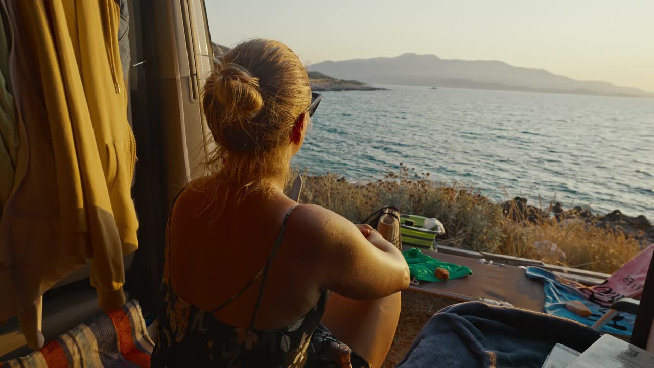 女人坐在露营车里欣赏美丽的海景。女人在海上看日落。有思想的女人在露营车上放松，欣赏日落时美丽的海景。视频素材