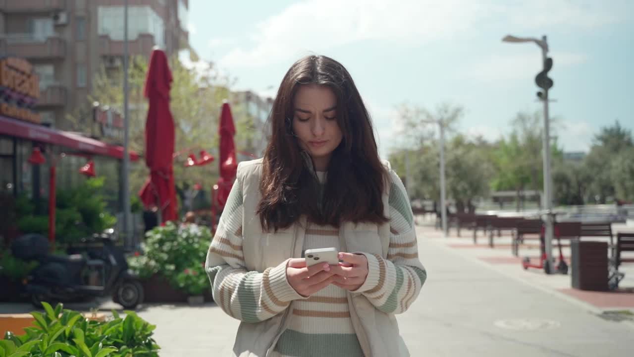 快乐的年轻女子在户外用手机打字。欢快的少女在城市背景下用智能手机散步。社交媒体，信使，暴徒应用，看视频，网上购物，浏览互联网。日常生活中的科技视频素材