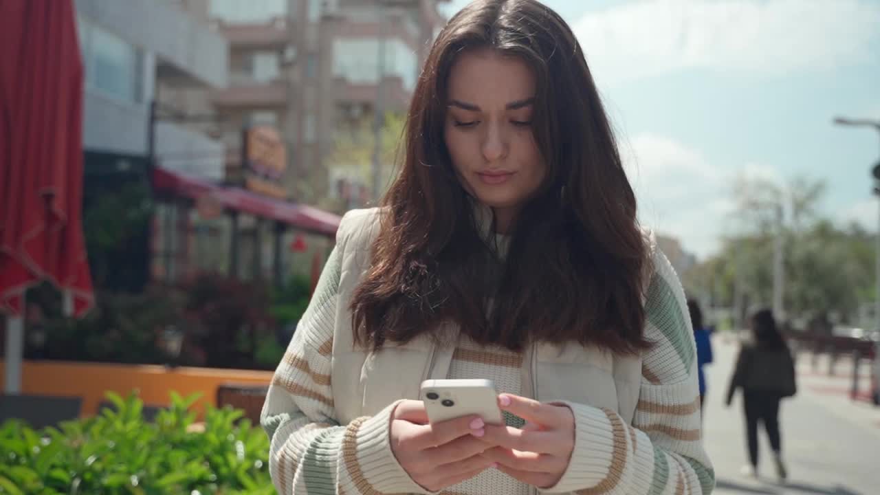 快乐的年轻女子在户外用手机打字。欢快的少女在城市背景下用智能手机散步。社交媒体，信使，暴徒应用，看视频，网上购物，浏览互联网。日常生活中的科技视频素材