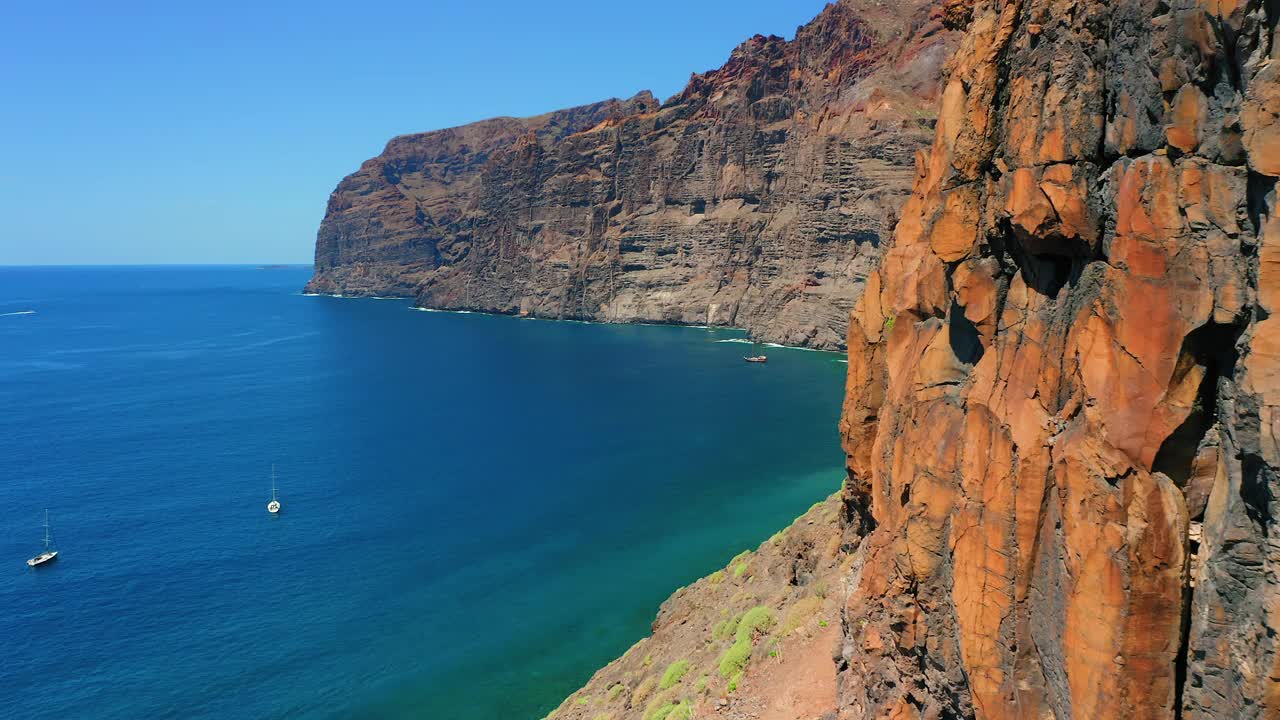 西班牙加那利群岛特内里费岛深海海岸线上的岩石火山巨大悬崖。湛蓝平静的海水，船在万里无云的天空。空中电影般的飞行。视频素材