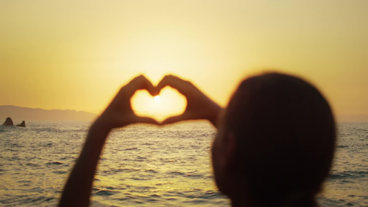 在海滩度假的女游客。夕阳下的女孩用手展示心形图案。夏季旅行者做梦。指缝间的阳光。视频素材