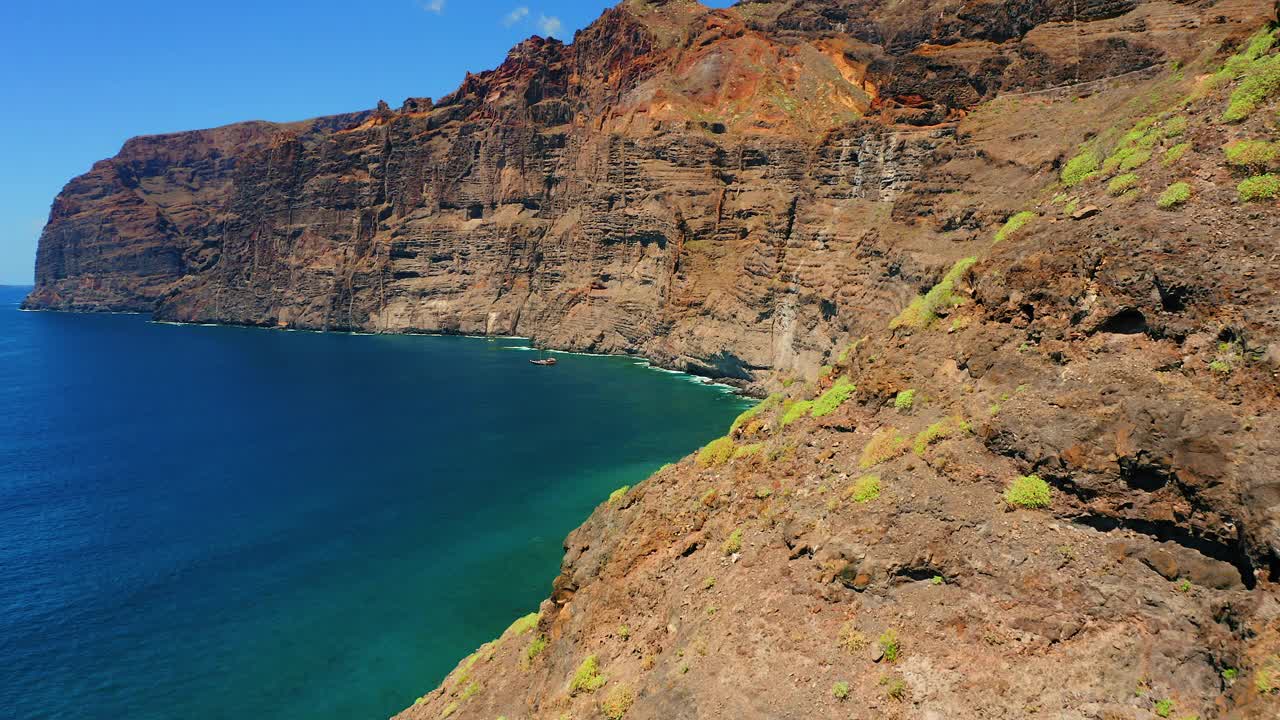 岩石火山巨大陡峭的悬崖与深海海岸线洛斯吉甘蒂斯特内里费加那利群岛。湛蓝平静的海水，船在万里无云的天空。空中电影般的飞行。视频素材