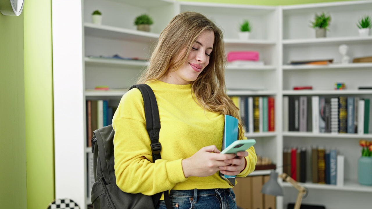 年轻的金发女学生在图书馆大学用智能手机拿着书视频素材