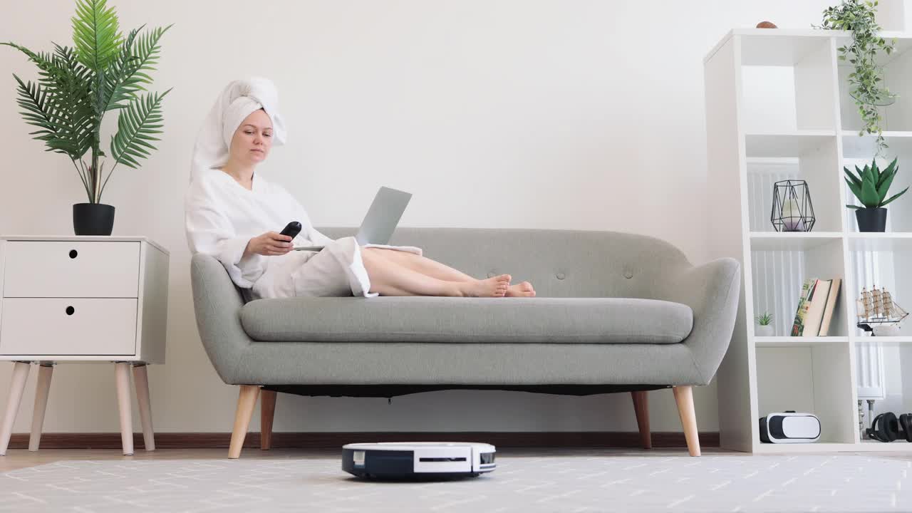 女人在沙发上拿着笔记本电脑休息，一边运行机器人吸尘器视频素材