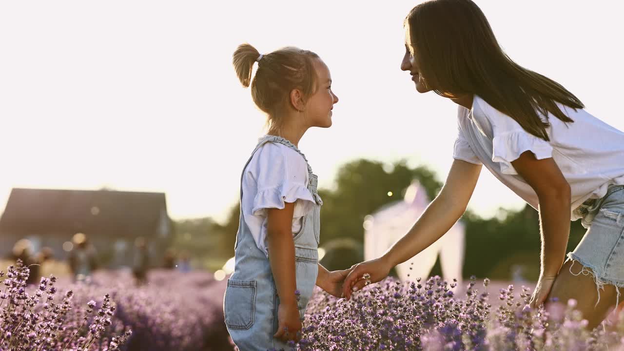 家庭日。夕阳下，年轻的妈妈和小女儿在薰衣草的田野里放松。一位美丽的母亲紧紧地拥抱并亲吻小女孩。母亲对孩子的关怀和爱。母亲节视频素材