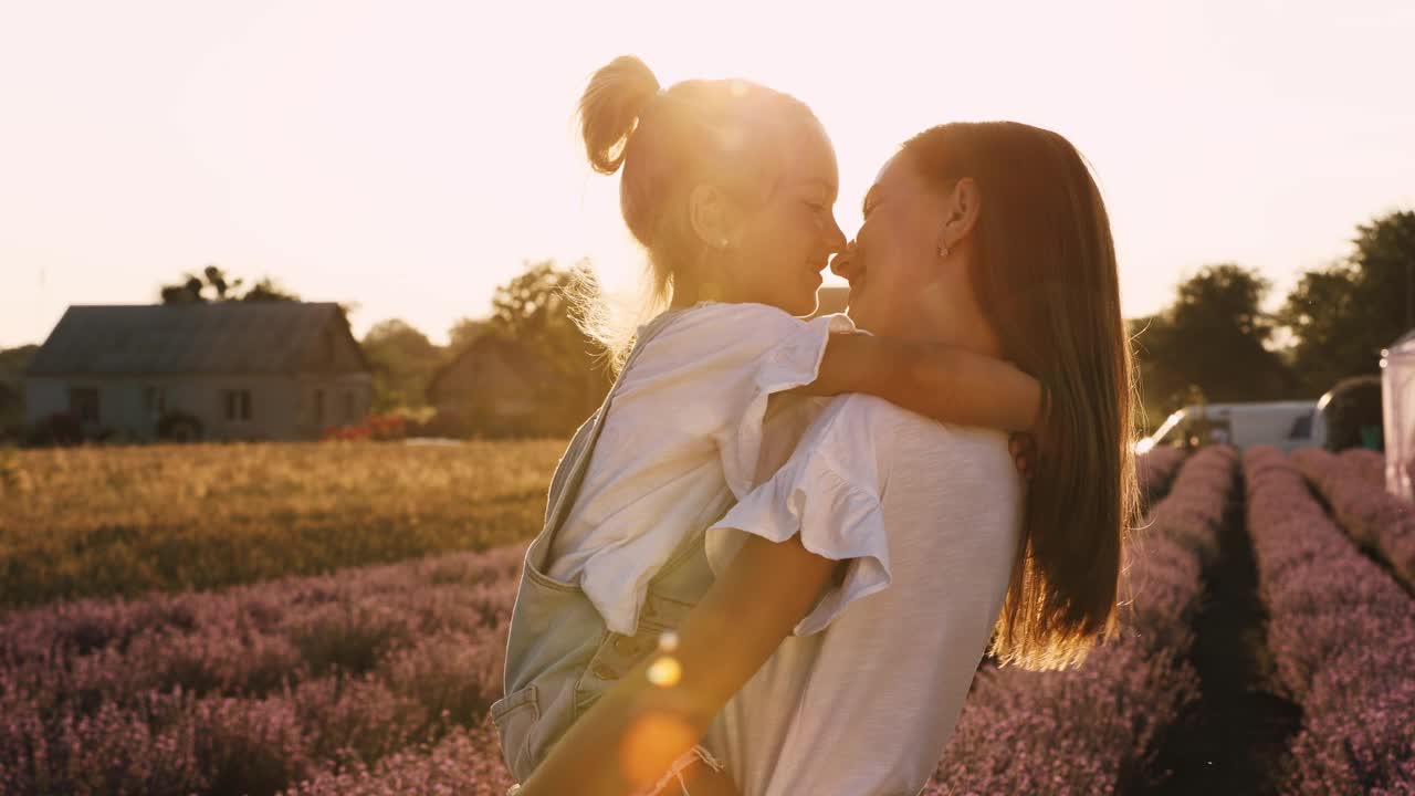 家庭日。夕阳下，年轻的妈妈和小女儿在薰衣草的田野里放松。一位美丽的母亲紧紧地拥抱并亲吻小女孩。母亲对孩子的关怀和爱。母亲节视频素材