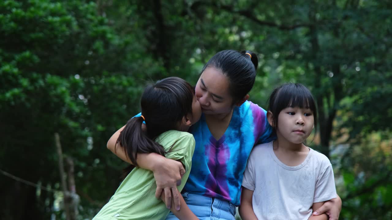 夏天，快乐的年轻母亲带着几个小女孩坐在河边的木桥上。母亲和两个女儿在户外玩得很开心。祝充满爱的家庭和母亲节快乐。视频素材