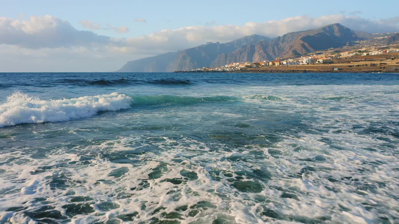 低低地飞过浪花翻滚的海面。盐水。度假小镇为背景，城市景观。岩石火山海滩在柔和的晨光特内里费岛。视频素材