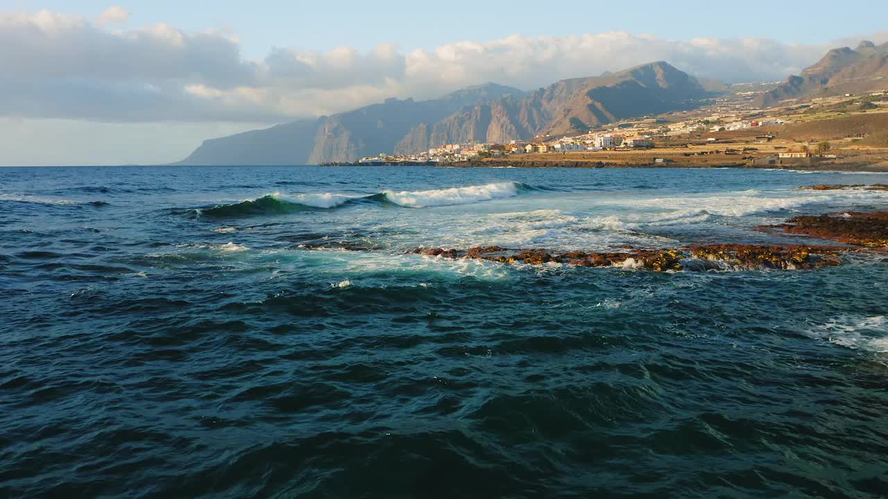 蓝色平静的海浪冲击着旅游城市附近的岩石海岸线，海滩上有火山岩。特内里费岛海岸的度假城市景观。西班牙的金丝雀。空中镜头。视频素材