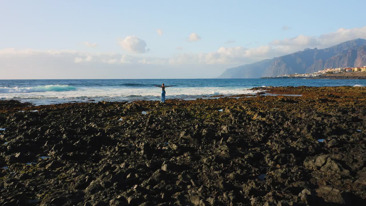 女孩旅行者举起她的手在岩石海滩与宽阔的泡沫海浪和巨大的悬崖为背景。女人喜欢海洋海岸线。旅游，旅游，度假在蓝色的大海。休闲。视频素材