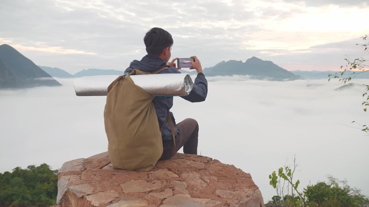 亚洲徒步旅行者男性坐在岩石上，用智能手机拍摄山顶雾山的照片视频素材