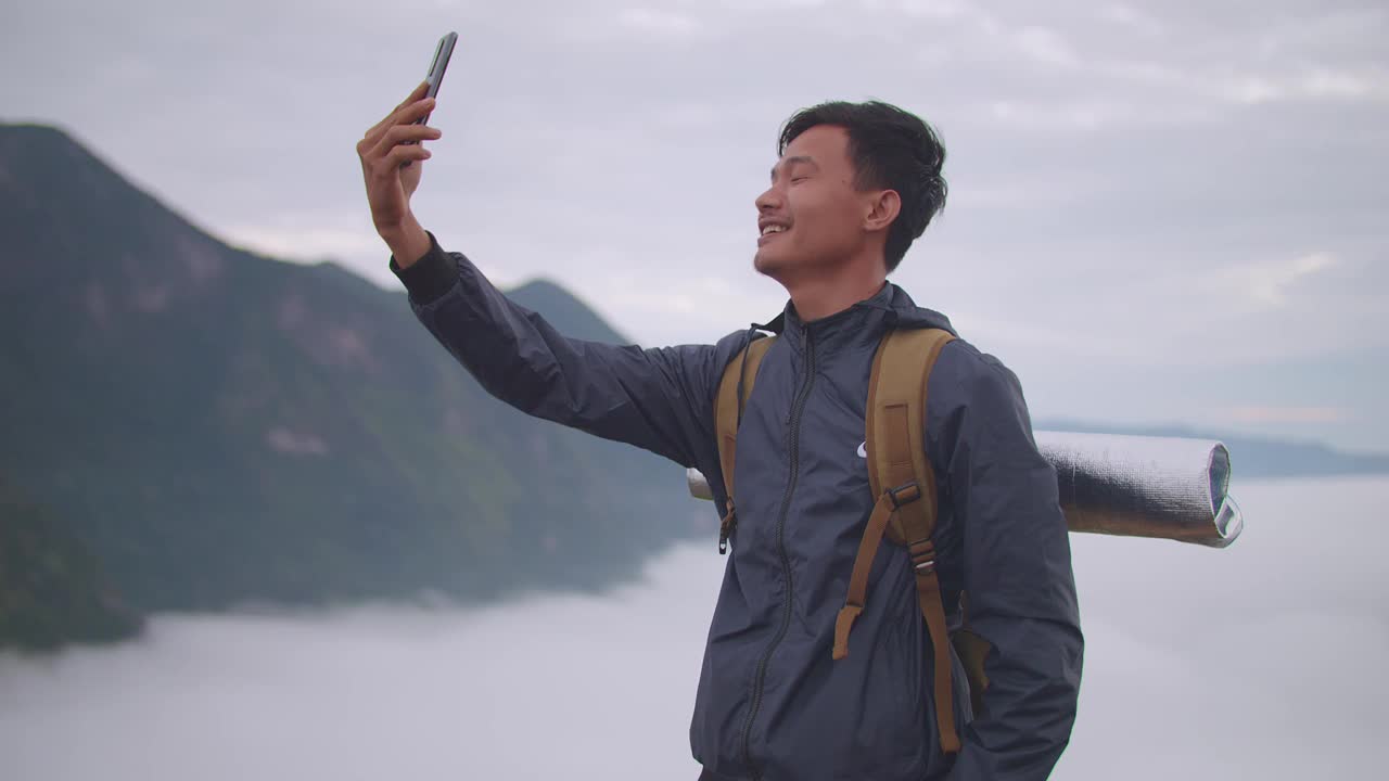 亚洲徒步旅行者男性微笑着对着智能手机说话，同时直播山顶雾山的景色视频素材