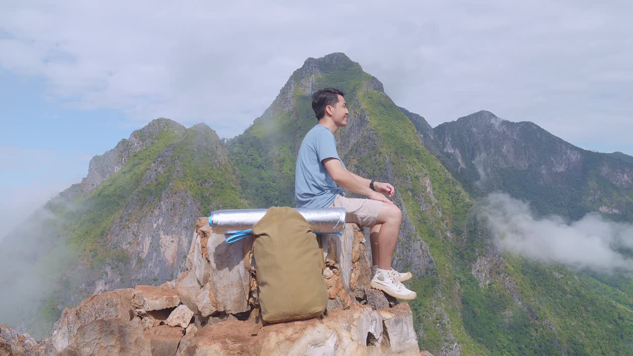 亚洲徒步男子背着背包坐在岩石上欣赏山顶雾山的美丽景色视频素材