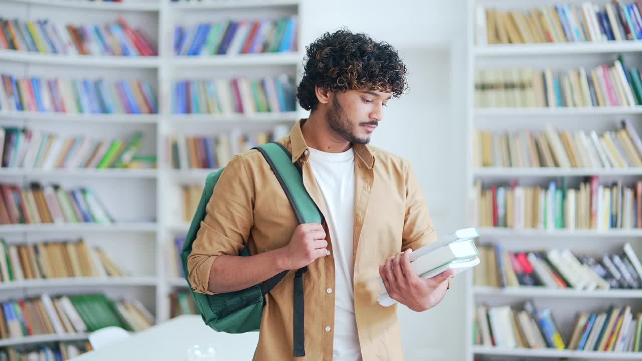 年轻的大学生微笑着拿着书，教科书，站在校园图书馆的空间里看着相机视频素材