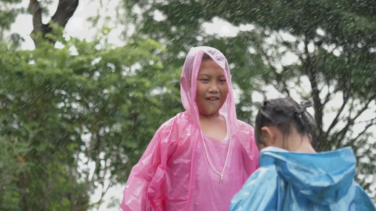 穿着雨衣的亚洲男孩带着美丽的微笑在雨中帮助他的小妹妹戴着头巾雨衣的慢动作。一个善良的孩子是未来可持续思考的人。视频下载