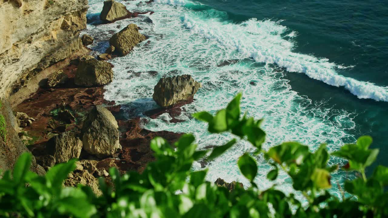 巴厘岛的绿色热带海岸线，石灰岩陡峭的悬崖和蔚蓝的绿松石般的印度洋水域。海浪泡沫。乌鲁瓦图岩石海滩。印度尼西亚。绿色植物的特写镜头。视频素材