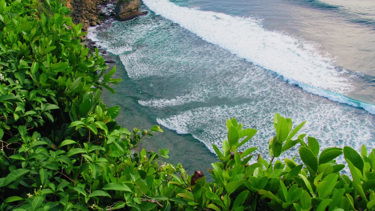 放松海景与绿松石宽泡沫浪滚在岩石海滩蓝色水晶清澈的水。宁静的禅意绿色自然背景。绿色植物，前景中的热带植物。视频素材