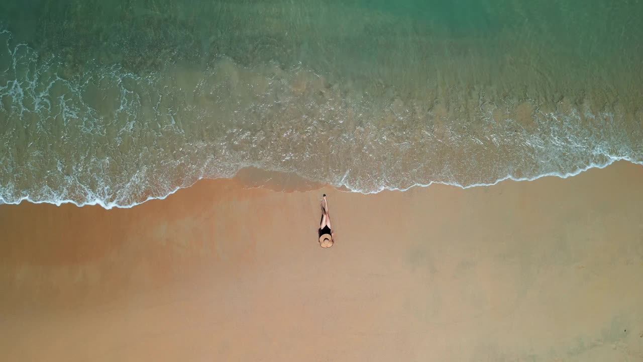 鸟瞰图，一名女子在有波浪的海滩上晒日光浴，她穿着泳衣遮住了脸。视频素材