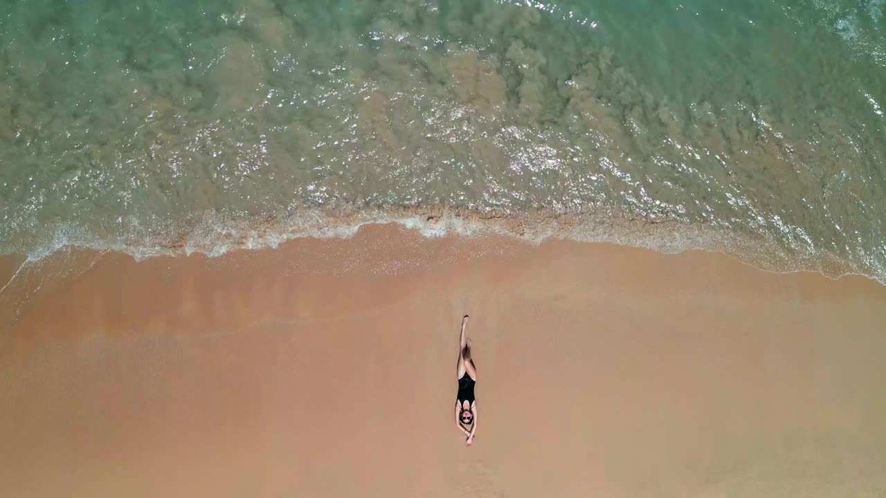 鸟瞰图:一名女子穿着泳衣在海滩上晒太阳，海浪拍打着她。视频素材