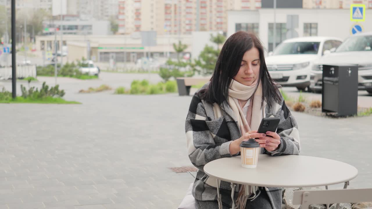 迷人而又讨人喜欢的年轻女子坐在街边咖啡馆的桌子旁，用纸杯喝咖啡，用智能手机浏览互联网和社交媒体视频素材