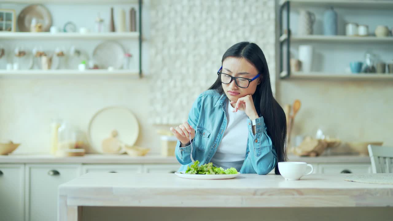 厌倦了节食减肥的年轻亚洲妇女坐在家里厨房的餐桌上，端着一盘沙拉视频素材