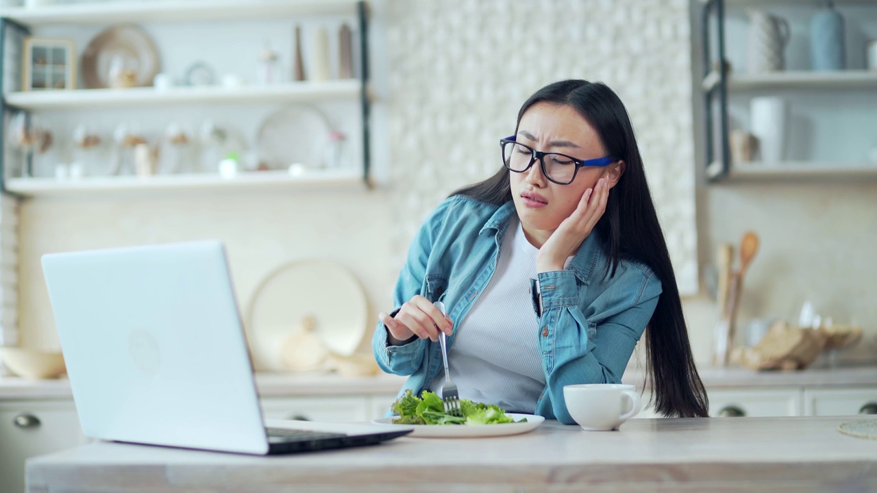 在家里的厨房里，一个疲惫的年轻亚洲女人坐在餐桌旁，端着一盘沙拉，看着笔记本电脑上的食谱视频素材