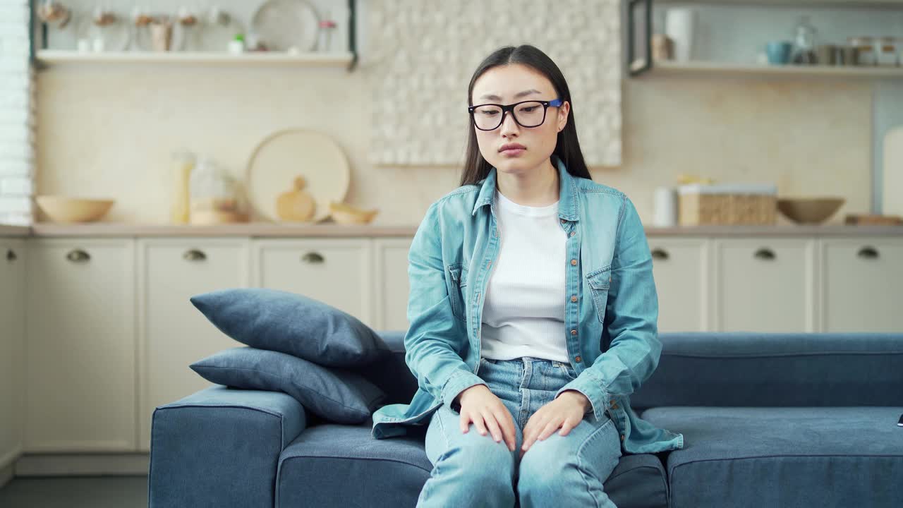 年轻的亚洲女子坐在客厅的沙发上感到胃紧张性疼痛。一名穿着便服的女性胃部不适，在家中患有急性胃炎视频素材