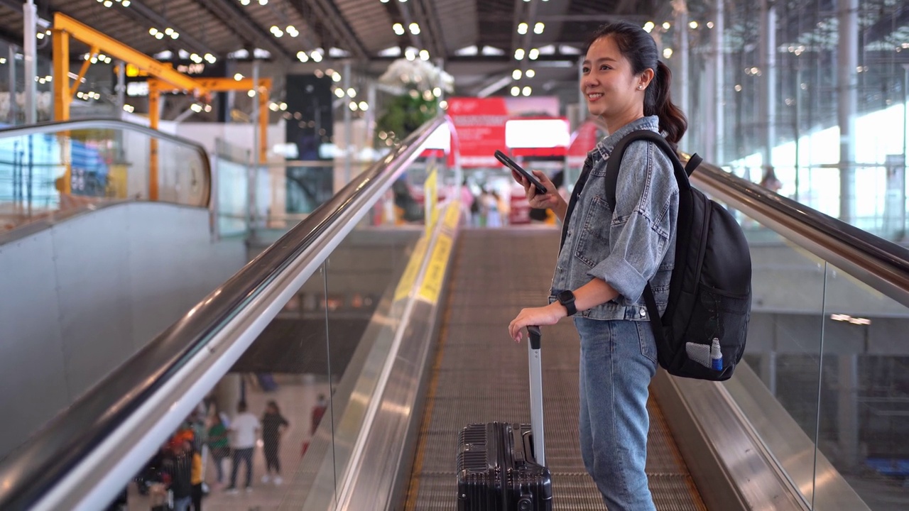 亚洲女性旅客在机场使用智能手机。她在使用自动扶梯时提着一个手提箱。视频素材