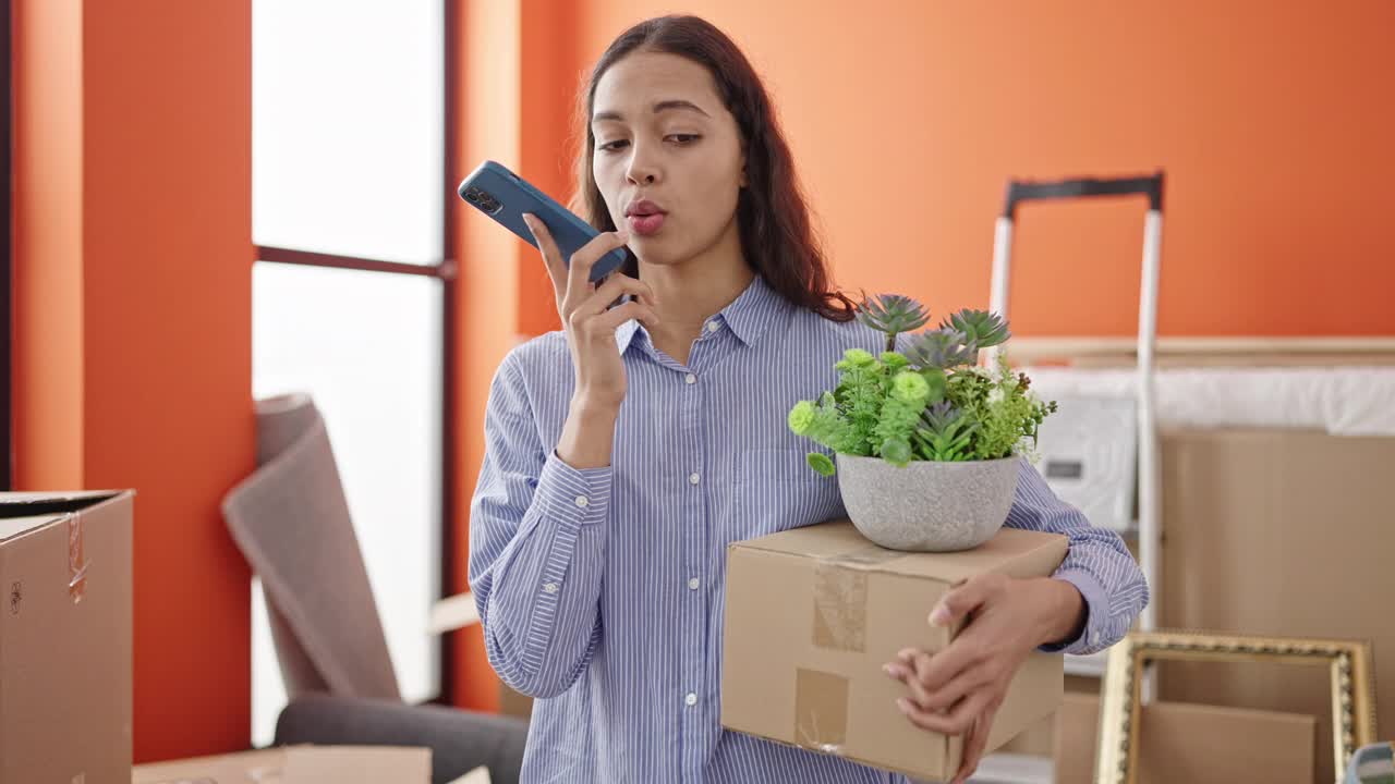 年轻美丽的西班牙女子在新家拿着盒子用智能手机发语音信息视频素材