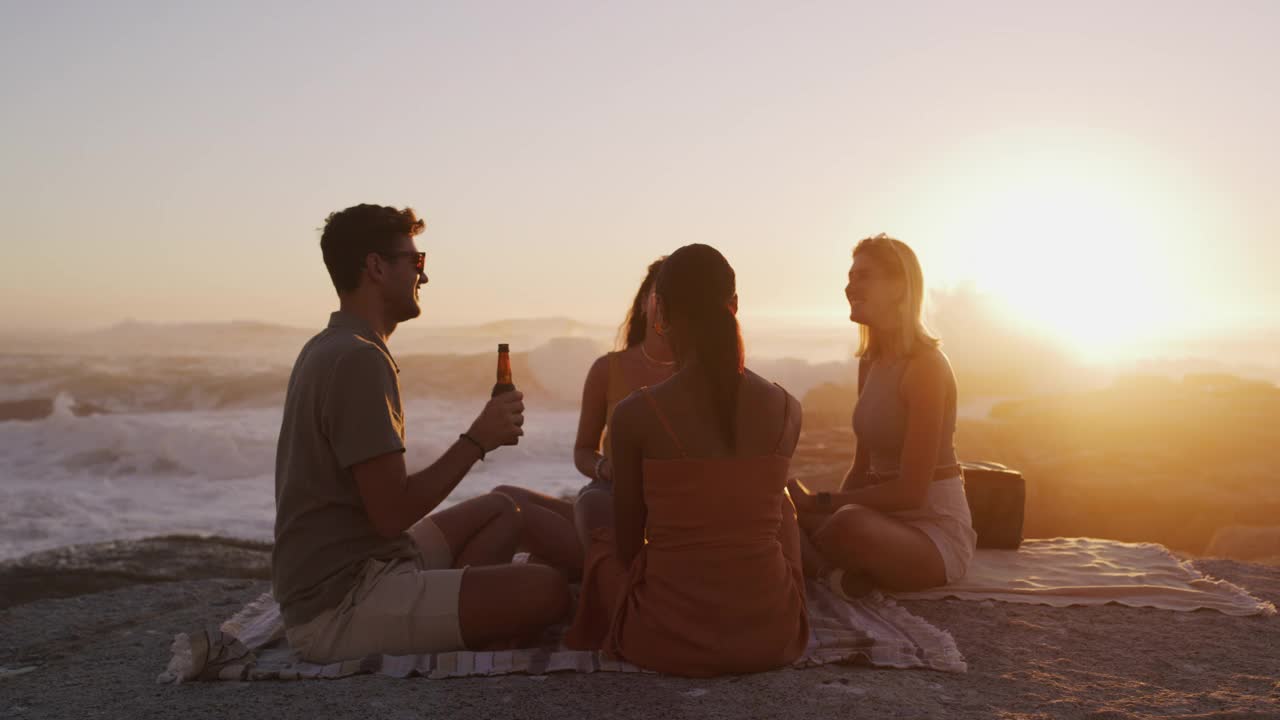 朋友们聚在一起，在日落的海滩上喝啤酒，一起野餐。男人和女人在户外分享吐司，庆祝自由旅行度假，假日或自然探险视频素材