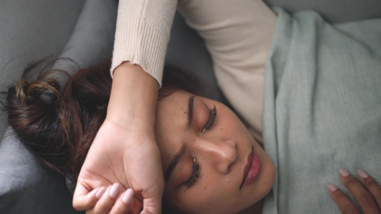 年轻的亚洲沮丧的女人躺在沙发上，她的手臂环绕着自己视频素材