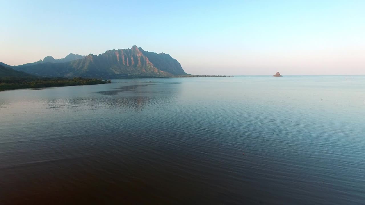 航拍日落时山脉附近的大海，无人机在绿色的草地和树木上向后上升-夏威夷瓦胡岛视频素材