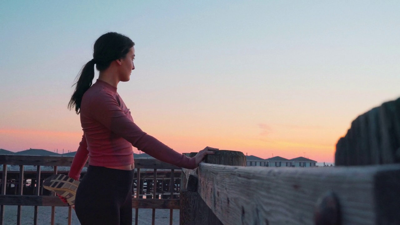 在加州圣地亚哥太平洋海滩的水晶码头上，一位年轻的慢跑女子在日落时分伸展身体，目光远离镜头视频素材